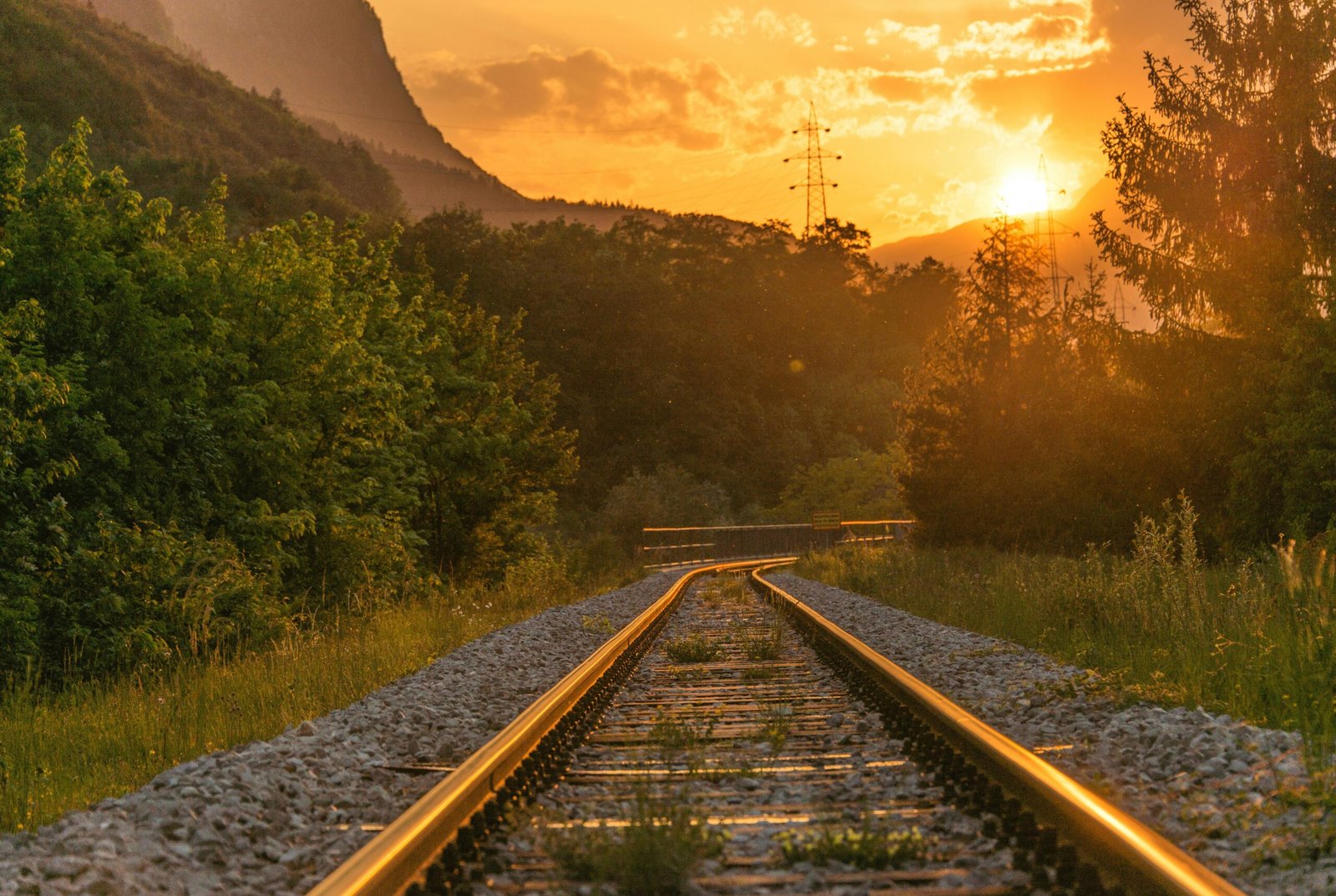 Golden sunset over railway tracks surrounded by lush greenery and mountains.